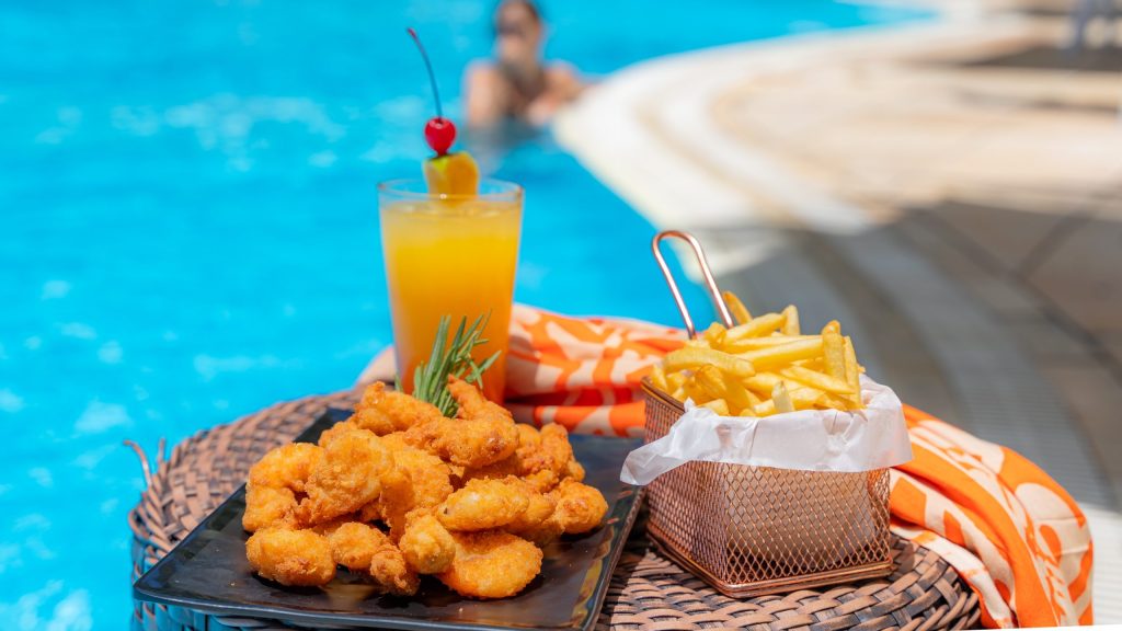 Mesa à beira da piscina com porção de frango empanado, batatas fritas em cestinha de metal e suco laranja decorado com frutas tropicais. Ao fundo, pessoa relaxa na piscina do Esmeralda Praia Hotel.
