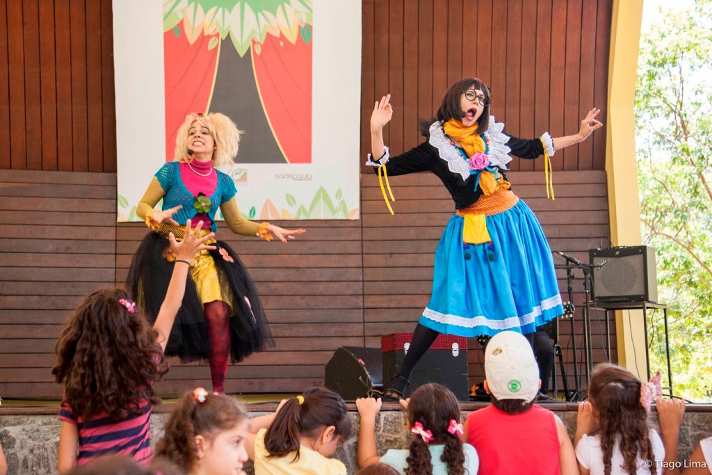 Duas mulheres apresentam-se em um palco ao ar livre; uma com peruca loira e roupa colorida, e outra com óculos e vestido tradicional, ambas expressando alegria e entusiasmo.