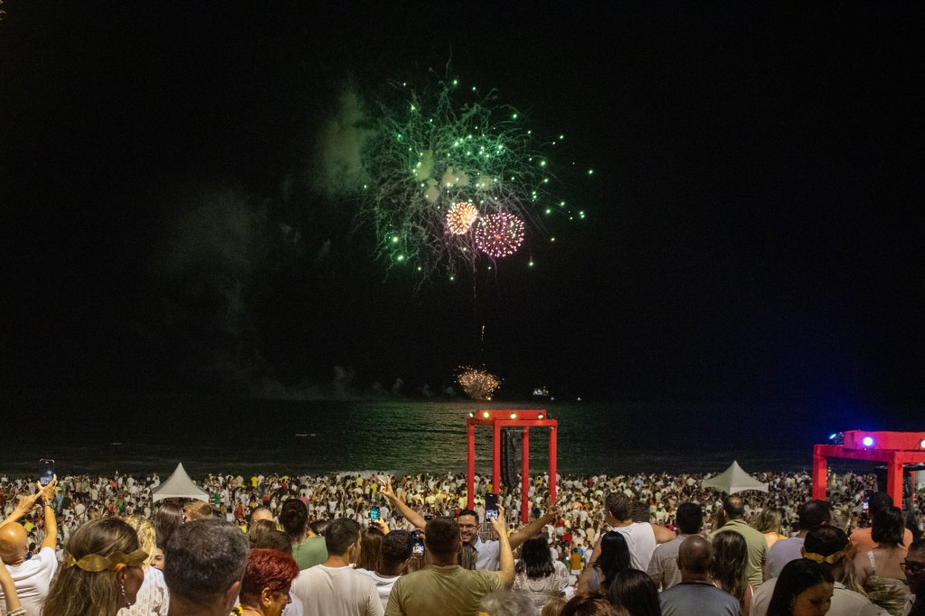 Multidão celebra o Réveillon na Praia de Ponta Negra, Natal-RN, assistindo a fogos de artifício coloridos sobre o mar à noite.
