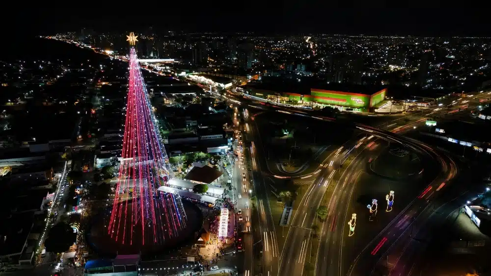 Vista aérea noturna da Árvore de Natal de Mirassol iluminada em Natal (RN), cercada por avenidas movimentadas e luzes da cidade ao fundo.