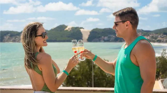 Casal no Esmeralda Praia Hotel brindando suas ta�as com champagne com o Morro do Careca ao fundo.