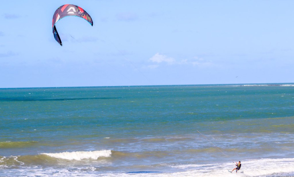 Pessoa praticando kitesurf na Praia de Ponta Negra, Natal-RN, com céu azul e mar em tons de verde e azul ao fundo.