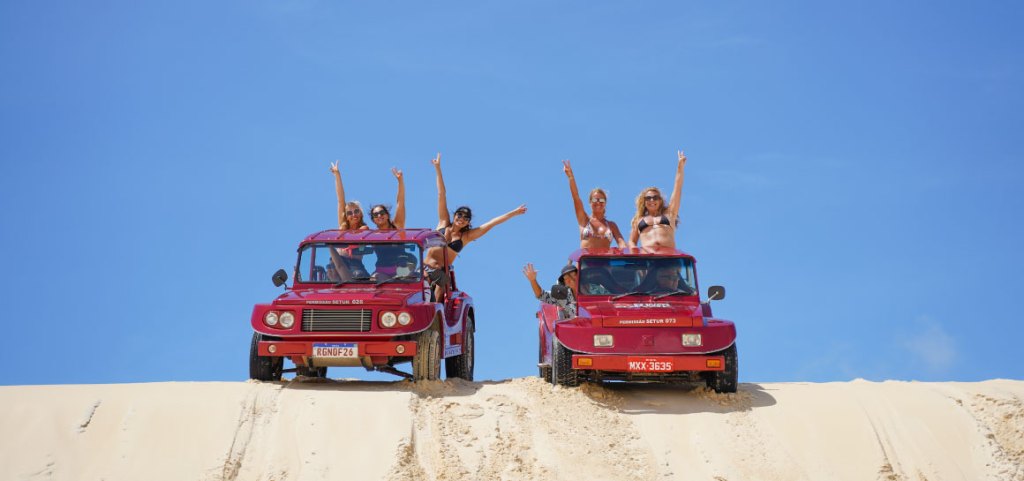 Grupo de pessoas em dois buggies vermelhos, comemorando no topo das dunas de Genipabu, Natal-RN, com céu azul ao fundo.