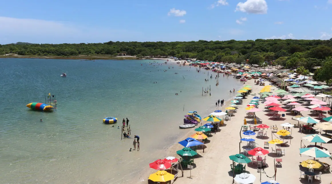 Vista aérea da lagoa de Carcará em um dia ensolarado, com uma praia cheia de guarda-sóis coloridos e visitantes desfrutando de atividades aquáticas.