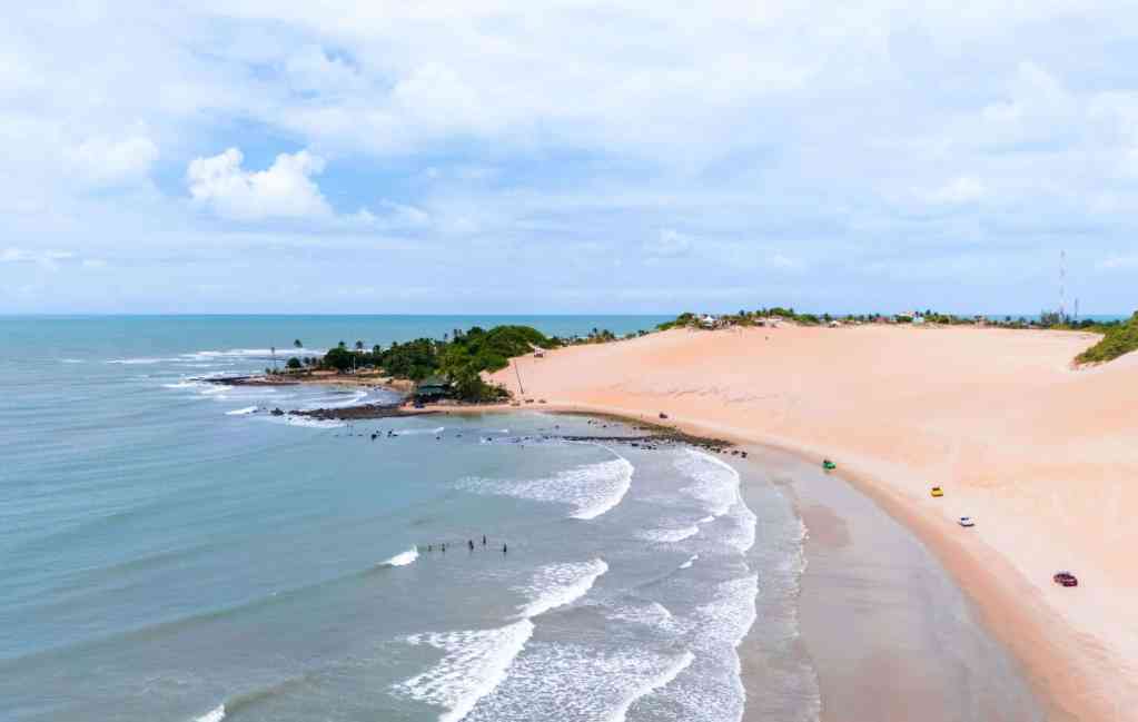 Imagem de drone mostrando a Praia de Genipabu com dunas extensas, mar calmo e buggy circulando na faixa de areia