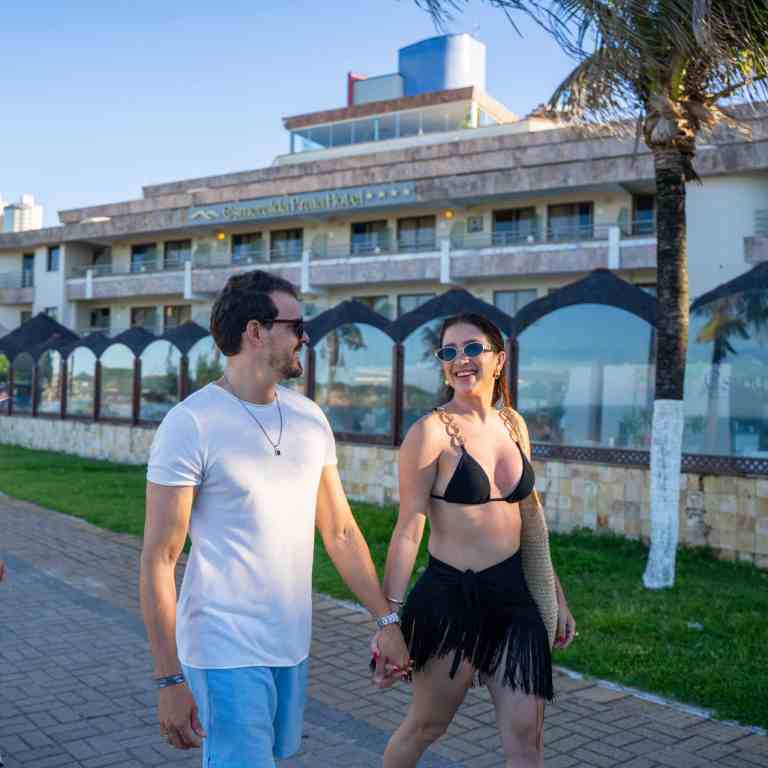 Hóspedes sorridentes de mãos dadas em passeio na orla de Ponta Negra com o Esmeralda Praia Hotel ao fundo em Natal RN