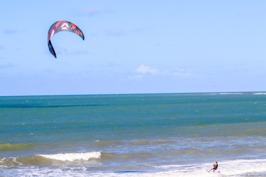 Pessoa praticando kitesurf no mar de Natal, conduzida por uma pipa colorida sob céu azul.