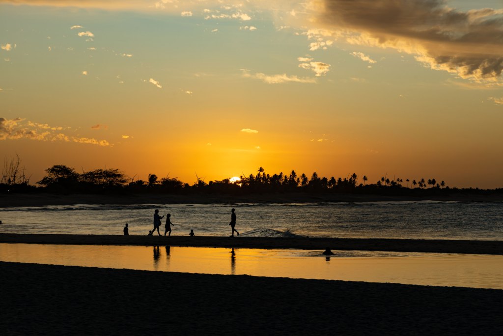 Silhuetas de pessoas caminhando e crianças brincando à beira-mar em São Miguel do Gostoso durante um pôr do sol dourado, com coqueiros ao fundo.