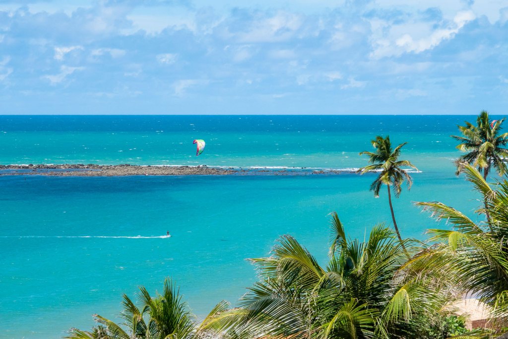 Windsurfista no mar azul próximo a recifes e coqueiros.