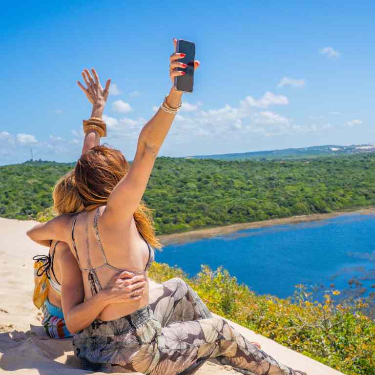 Duas-turistas-abracadas-registrando-momento-em-duna-de-areia-com-paisagem-panoramica-da-lagoa-e-vegetacao-tropical-em-Genipabu-Natal-RN.jpg
