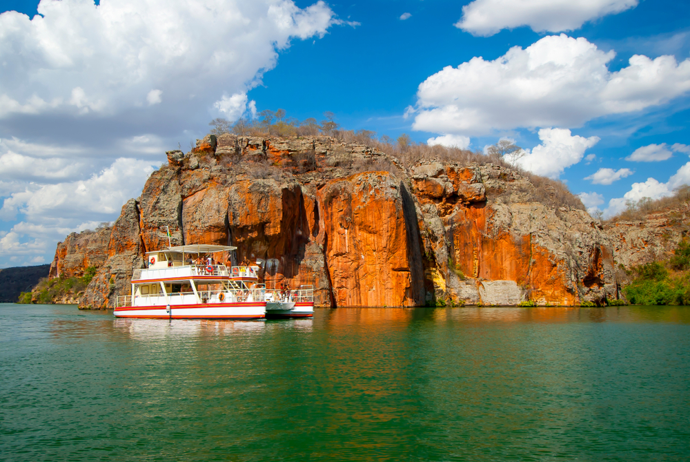 Barco turístico navega em águas verdes diante do Canyon do Xingó, Sergipe, Brasil, sob céu azul com nuvens brancas, em cenário natural deslumbrante.