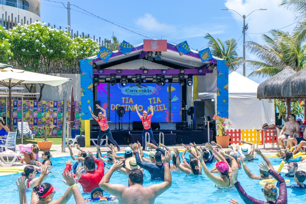Foto da piscina do Esmeralda Praia Hotel com hóspedes com os braços para cima curtindo a hidroginástica. Equipe de recreação e palco com decorações temáticas ao fundo.
