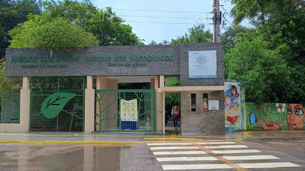 Portão de entrada do Parque das Dunas Jornalista Luiz Maria Alves, no Bosque dos Namorados, em Natal (RN), com fachada de pedra, grades verdes e murais coloridos ao lado.