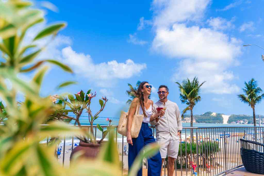 Casal sorridente no deck suspenso do Esmeralda Praia Hotel, segurando drinks, com vista para o mar, coqueiros e o Morro do Careca.