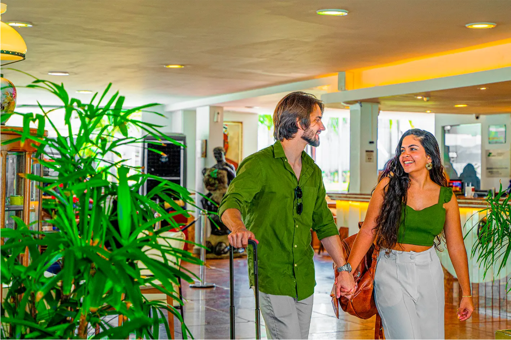 Casal sorridente caminha de mãos dadas pelo lobby do Esmeralda Praia Hotel, carregando malas de viagem, cercado por plantas e decoração do saguão